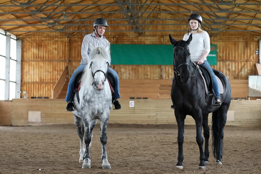 two horse and rider pairs stand in an indoor arena