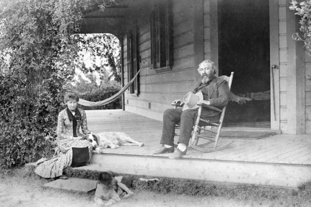 black and white photo of man holding hat in rocking chair on porch with woman and two dogs